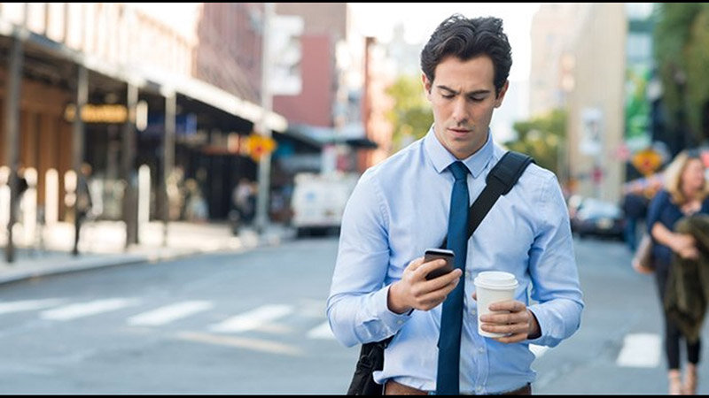 Image: A businessman wearing a blue shirt and tie, walking on a city street, holding a coffee cup and looking at cell phone.